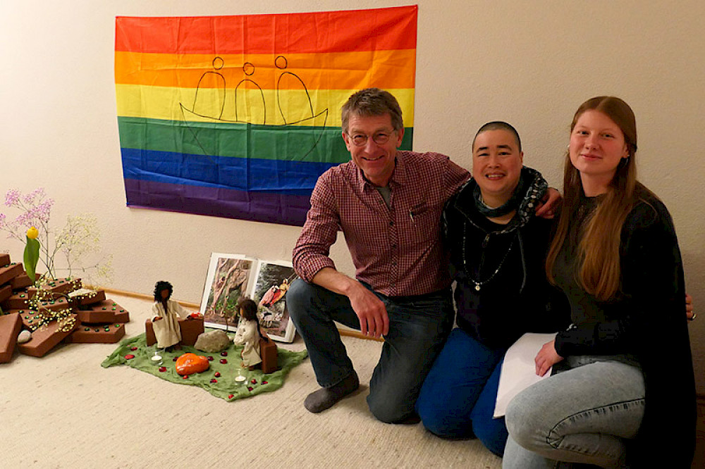 Bild zeigt drei Personen vor einer Regenbogenflagge mit dem Arche-Logo.